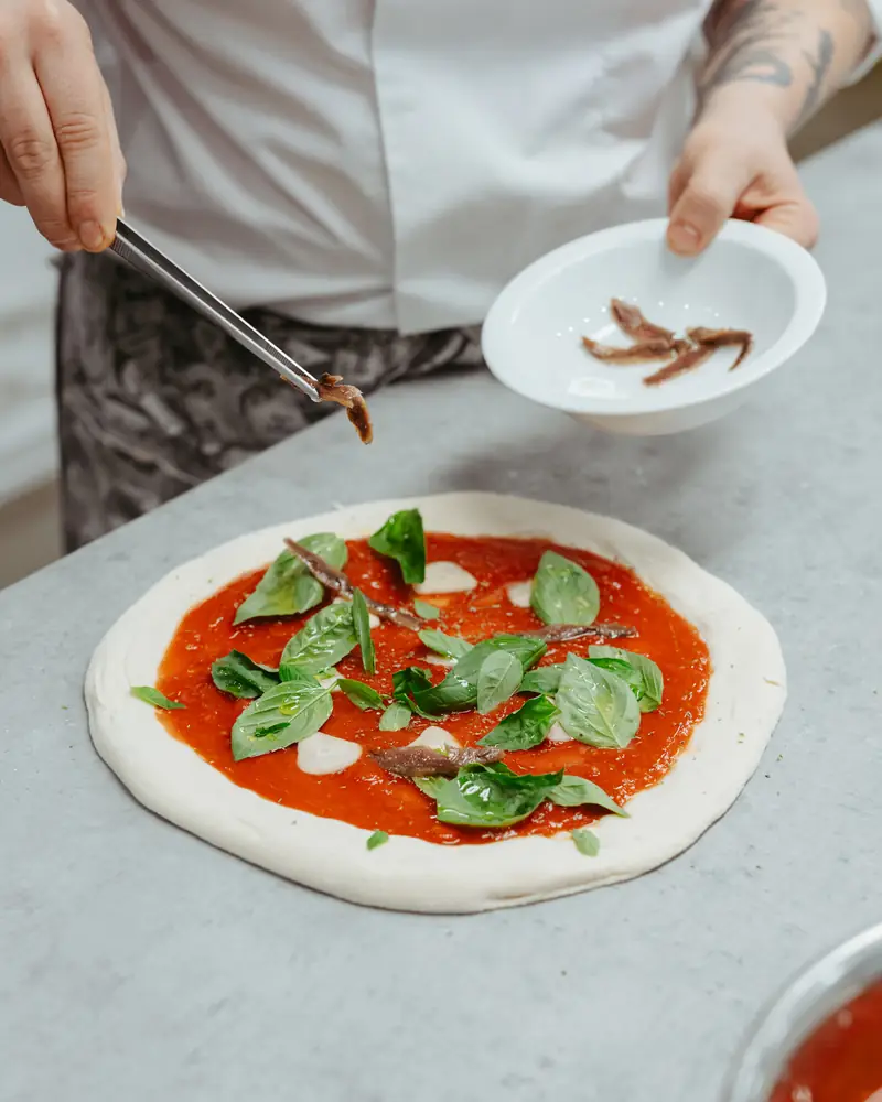 Dłonie pizzaiolo układają bazylię na pizzy przed wypiekiem
